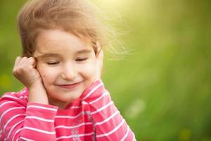 Little girl in a coral striped T-shirt on a green background in a field holds her face in her hands and smiles slyly. Children's Day, happy child, environmental and nature protection, insect repellent photo