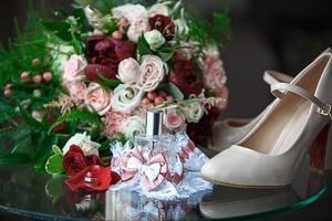 accesorios de boda para la novia y el novio un ramo de rosas rojas, rosadas y blancas, un boutonniere, anillos de boda de oro, una liga de encaje con un lazo, perfume. zapatos beiges joyas, foto