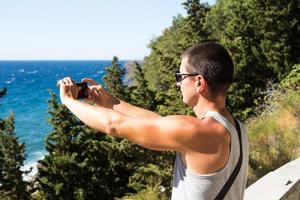 Male tourist takes photos on a smartphone of beautiful view of sea in a mountainous area from the top among coniferous trees. Video communication, using the phone when traveling. SIM card in roaming