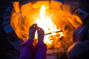 Bare feet of child by fire. Gatherings at night by campfire in open air in summer in nature. Family camping trip, gatherings around the campfire. Camping lantern and tent. Warm your feet, cold night