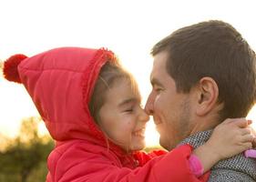 Little girl in red jacket with a hood hugs and kisses her dad, smiles, touches her nose. Happy family, children's emotions, father's day, bright rays of the sun, Caucasian appearance. Space for text. photo