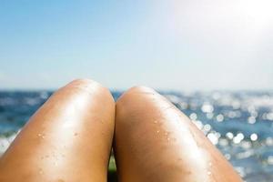 Smooth tanned female legs in the spray of water on the beach against the background of the sea. Tanning products, sunburn protection, skin care, depilation, beach holidays. Copy space photo