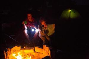 papá e hija se sientan por la noche junto al fuego al aire libre en verano en la naturaleza. viaje de campamento familiar, reuniones alrededor de la fogata. día del padre, barbacoa. linterna de camping y carpa foto