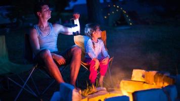 Dad and daughter sit at night by the fire in the open air in the summer in nature. Family camping trip, gatherings around the campfire. Father's Day, barbecue. Camping lantern and tent photo