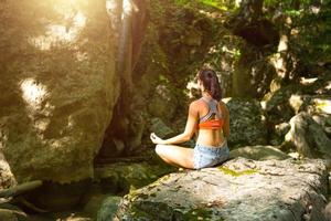 una mujer se sienta en un campo de loto sobre una gran roca entre las rocas al aire libre y medita, disfruta de la unidad con la naturaleza, escucha el silencio y los sonidos del bosque. ecología