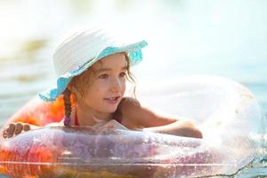 chica con sombrero nadando en el río con un círculo inflable transparente en forma de corazón con plumas naranjas dentro.el mar con un fondo arenoso. vacaciones en la playa, nadar, broncearse, protectores solares. foto