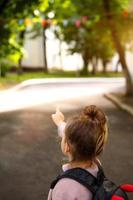 A little girl of Caucasian in a school uniform with a backpack looks at the road in the school yard. Concept back to school. Elementary school, developing activities for preschoolers. Space for text photo