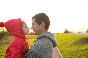 Little girl in red jacket with a hood hugs and kisses her dad, smiles, touches her nose. Happy family, children's emotions, father's day, bright rays of the sun, Caucasian appearance. Space for text.
