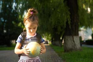 Kaluga, Russia-June 23, 2020. A girl from an elementary school of Caucasian appearance with a backpack on her shoulders holds a globe in her hands and carefully studies it. photo