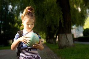 una niña de una escuela primaria de apariencia caucásica con una mochila en los hombros sostiene un globo en sus manos y lo estudia cuidadosamente. educación, regreso a la escuela, 1 de septiembre. espacio de copia