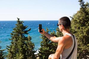 Male tourist takes photos on a smartphone of beautiful view of sea in a mountainous area from the top among coniferous trees. Video communication, using the phone when traveling. SIM card in roaming