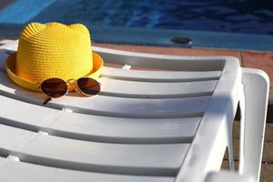 Beach accessories a yellow hat and sunglasses lie on a white deck chair by the pool. Sun protection, UV, tan, travel. Vacation concept, sea trip, swimming. Copy space. Flatlay photo