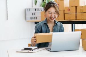 pequeña empresa emprendedora de mujeres asiáticas jóvenes que trabajan con una computadora portátil para comprar en línea en casa, alegre y feliz con una caja para empacar en casa, un negocio propio que comienza un negocio en línea