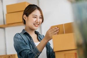 una mujer asiática inicia una pequeña empresa propietaria de una caja de cartón en el lugar de trabajo. una vendedora independiente prepara una caja de paquetes de productos para entregar al cliente. venta en línea, comercio electrónico foto