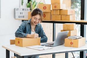 Asian woman entrepreneur is packing product to the box at home workplace. teenager business owner work at home, many stuff to do today in home. Start up SME concept. photo