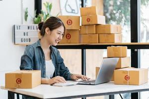 pequeña empresa emprendedora de mujeres asiáticas jóvenes que trabajan con una computadora portátil para comprar en línea en casa, alegre y feliz con una caja para empacar en casa, un negocio propio que comienza un negocio en línea