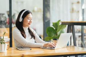 Smiling woman wearing wireless headphones working typing on notebook sit at desk in office workplace. Enjoy e-learning process, easy comfortable application usage, listen music during workday concept photo