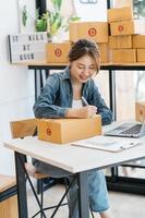 Asian woman entrepreneur is packing product to the box at home workplace. teenager business owner work at home, many stuff to do today in home. Start up SME concept. photo