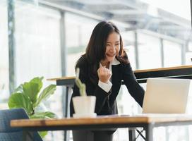 Excited young woman standing at table with laptop and celebrating success