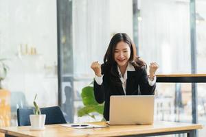 Excited young woman standing at table with laptop and celebrating success photo