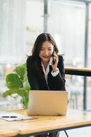 Happy smiling Asian business woman working on laptop at office, using smart phone. Businesswoman sitting at her working place photo