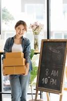 una mujer asiática inicia una pequeña empresa propietaria de una caja de cartón en el lugar de trabajo. una vendedora independiente prepara una caja de paquetes de productos para entregar al cliente. venta en línea, comercio electrónico. foto