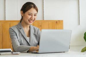 feliz joven mujer asiática sentada y usando una computadora portátil en la sala de estar trabajando desde el concepto de casa foto