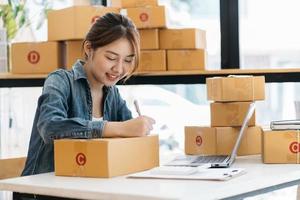una mujer asiática inicia una pequeña empresa propietaria de una caja de cartón en el lugar de trabajo. una vendedora independiente prepara una caja de paquetes de productos para entregar al cliente. venta en línea, comercio electrónico foto