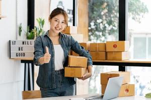 Asian woman entrepreneur is packing product to the box at home workplace. teenager business owner work at home, many stuff to do today in home. Start up SME concept. photo