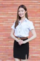 Portrait of an adult Thai student in university student uniform. Asian beautiful girl standing with her arms crossed on a brick background. photo