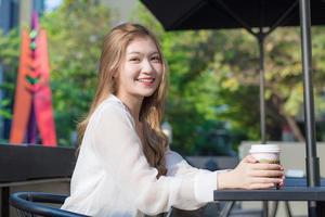 una joven mujer asiática hermosa que usa traje con cabello de bronce se sienta sonriente en una silla en una cafetería al aire libre mientras sostiene una taza de café en su mano en una mañana soleada. foto