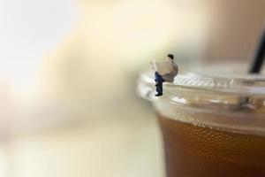 negocio, concepto de relajación. primer plano de la figura en miniatura de un hombre de negocios sentado y leyendo el periódico en una taza de plástico para llevar de café negro helado con espacio para copiar. foto