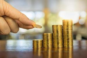 concepto de negocio, dinero, finanzas y ahorro. cierre la mano del hombre sosteniendo y ponga una moneda en la pila de monedas en la mesa de madera. foto