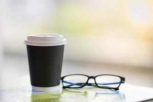 Close up of take away paper cup of hot coffee with reading glasses on world map with copy space. photo