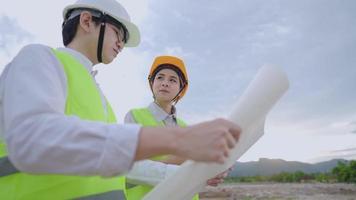 Asian young Engineer architecture wear safety working gear discussing on project at cloudy outdoor construction site, safety vest and safety helmet, brainstorm teamwork spirit working environment video