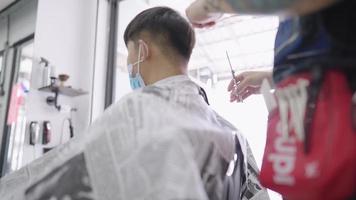 l'uomo asiatico indossa una maschera protettiva si taglia i capelli durante la pandemia di quarantena, lama per tosatrice, abilità di taglio delle forbici, riapertura del barbiere maschio durante la pandemia, prevenzione delle malattie, opportunità per piccole imprese video