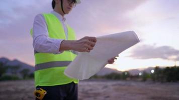 Portrait asian male engineer in protective yellow vest and hardhat, looking at paper blueprint inside puddle construction land, hand holding building plan contemplating on project, late working worker video