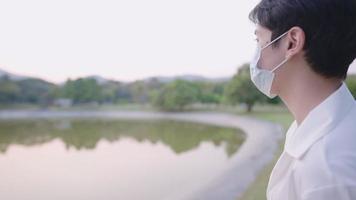 A young attractive asian man wears protective face mask standing next to a natural pond in city park, enjoys looking around a crowded park in evening, with people playing badminton behind, new normal video