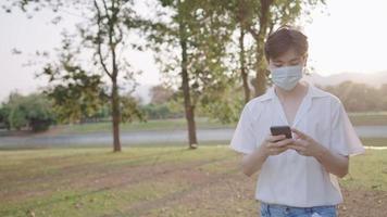 los jóvenes asiáticos usan máscaras faciales se conectan en línea usando un teléfono inteligente relajándose dentro del parque durante la puesta de sol, reflejo de bengalas de luz solar cálida, estilo de vida moderno, pandemia de bloqueo del parque al aire libre al aire libre video