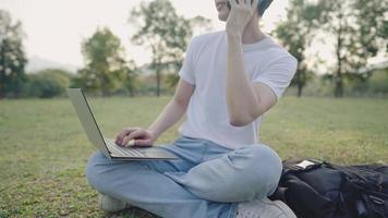 Young attractive asian multitasking freelancer sitting on a wide meadow, busy working outside on a portable laptop while talking on phone during day, distant communication technology and lifestyle video