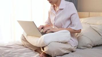 A young asian woman working with her laptop on the pillow sitting on the bed with warm morning light, work from home inside comfortable cozy bedroom, home wireless internet connection, shopping online video