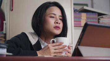 Asian woman drinking hot beverage, holding white coffee cup, watch tablet on relaxing night at home, study at home learning online wireless technology, digital media learning online entertainment video