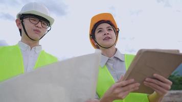 Mixed race construction workers, assistant in safety protective wear talking discussing into work project at construction land site, holding tablet blueprint paper, male and female engineer employee video