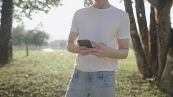 feche uma parte do corpo masculino na camisa branca usando smartphone enquanto caminhava sob a sombra das árvores dentro do parque público, atividade de lazer relaxante, dispositivos de comunicação distantes, mãos usam tecnologia sem fio video