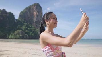 young beautiful asian woman video calling with friends through smartphone on Relaxing trip at the tropical island beach, wireless technology, lady waving to the phone camera showing enjoyment emotion