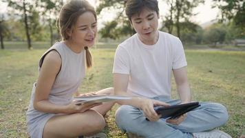 Young adult male and female friends sit down on the ground inside the park, discussing homework project, friendship socialize, doing group research thesis, look up information on tablet, outdoor scene