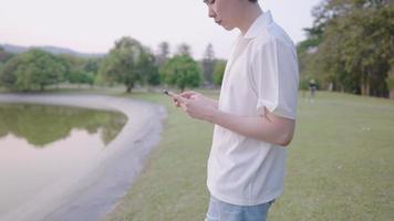 attraktiver, gut aussehender asiatischer mann, der tagsüber auf einer wiese im grünen park steht, remote an einem inspirierenden grünen ort arbeitet und das smartphone in der hand hält, um soziale anwendungen und technologie zu überprüfen video