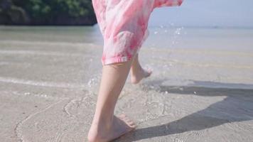 View from behind of female barefoot walking on the island beach, enjoying summer vacation. Hot sunny weather, walk in to clear ocean water, wave splashing slow motion, Freedom life escape from city