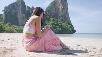 asiatische junge schlanke frau, die sich entspannt am telefon spielt, während sie am weißen strand an der wunderschönen asiatischen küste sitzt, die mobile app nutzt und ein sonnenbad macht, weibliche soloreise, remote-nutzung des drahtlosen internets video