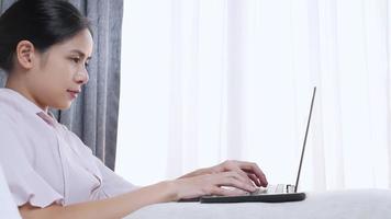 Asian woman working with laptop placing on lap on comfortable bed, work from home isolation, freelance author writing email to clients, fresh ideas in the morning, good work environment, morning light video